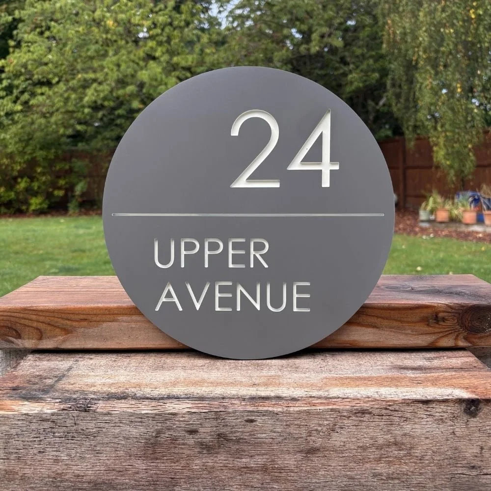 Round house number sign with '24 Upper Avenue' on a wooden surface outdoors.
