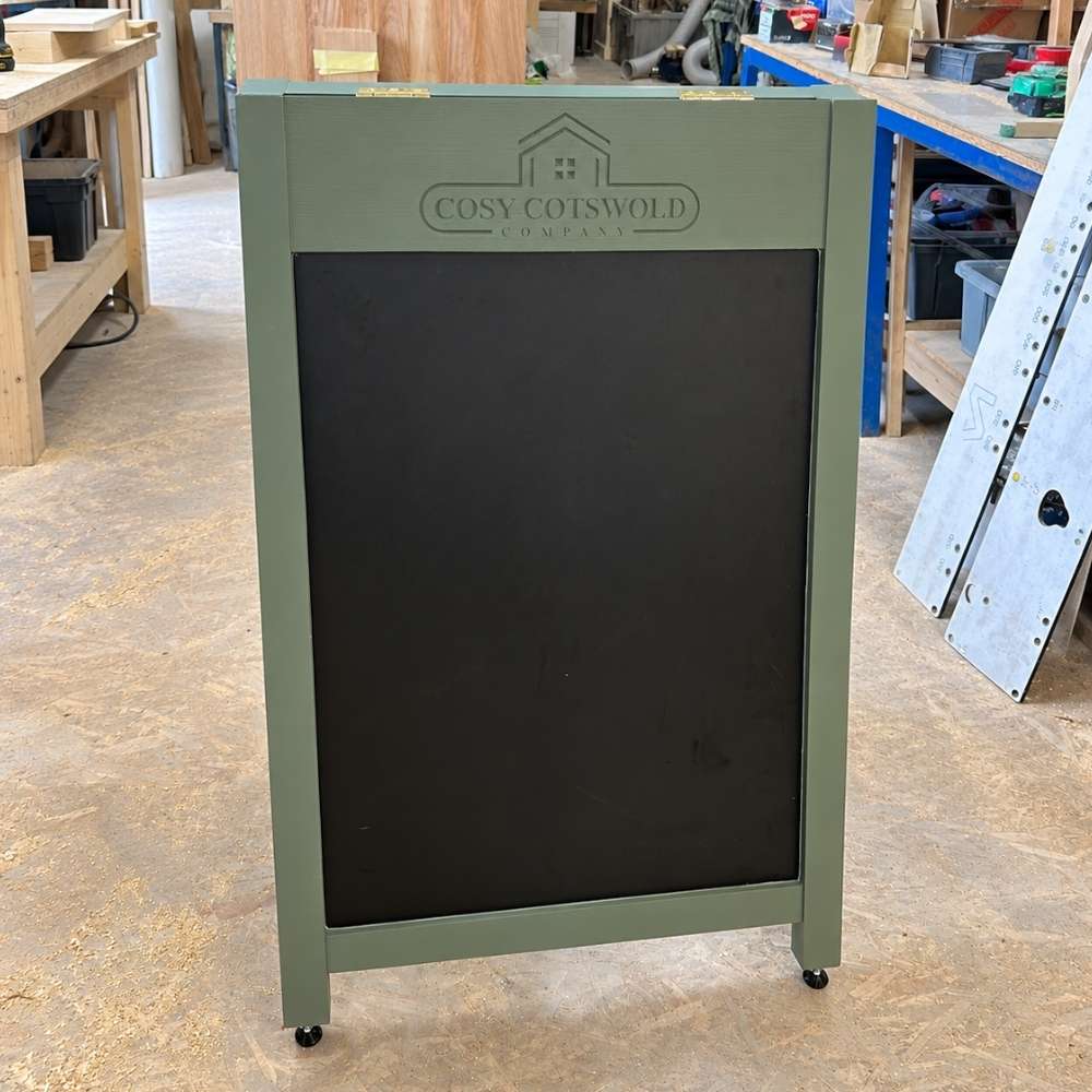 A freestanding A-frame chalkboard sign with an cotswold green frame and black writing surface, shown in a workshop. The top frame is embossed with the brand name 'COSY COTSWOLD COMPANY'.