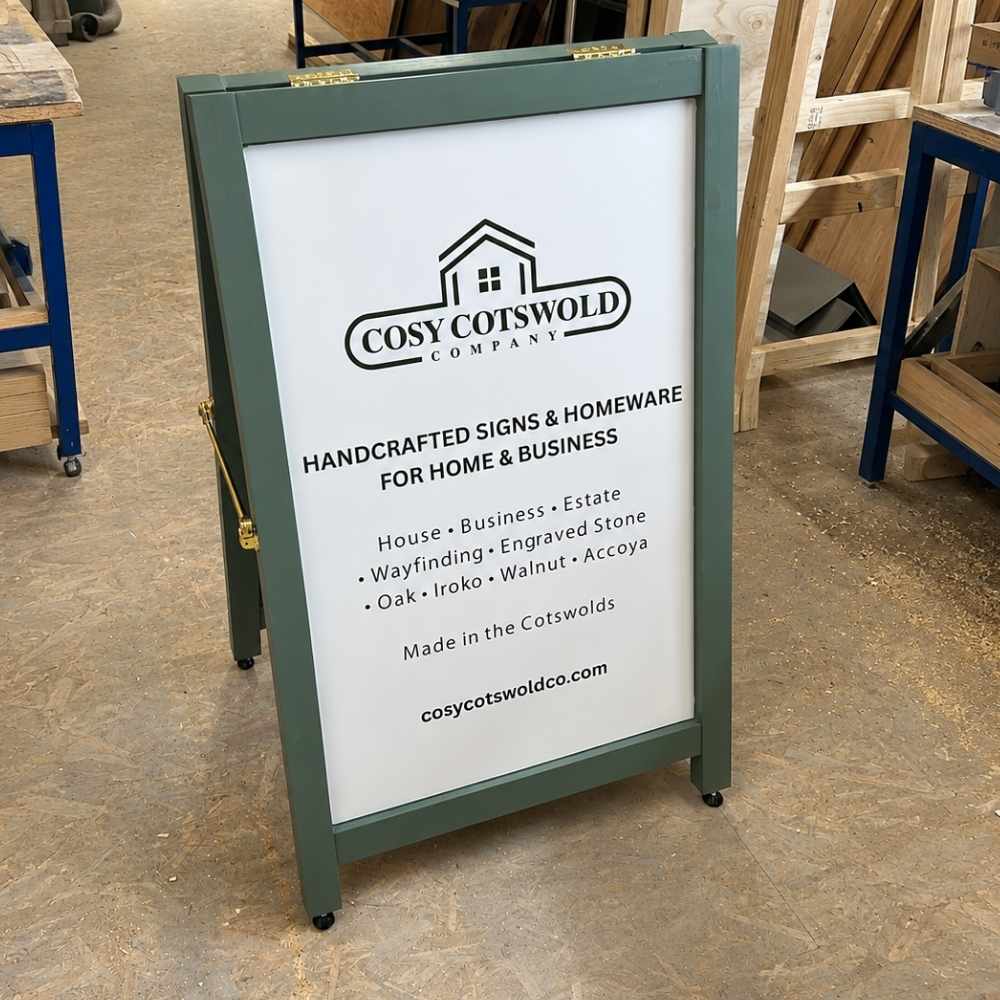 A freestanding A-frame sidewalk sign with a white panel and cotswold green frame, shown in a workshop setting. The sign features the Cosy Cotswold Company logo and text advertising handcrafted signs and homeware.