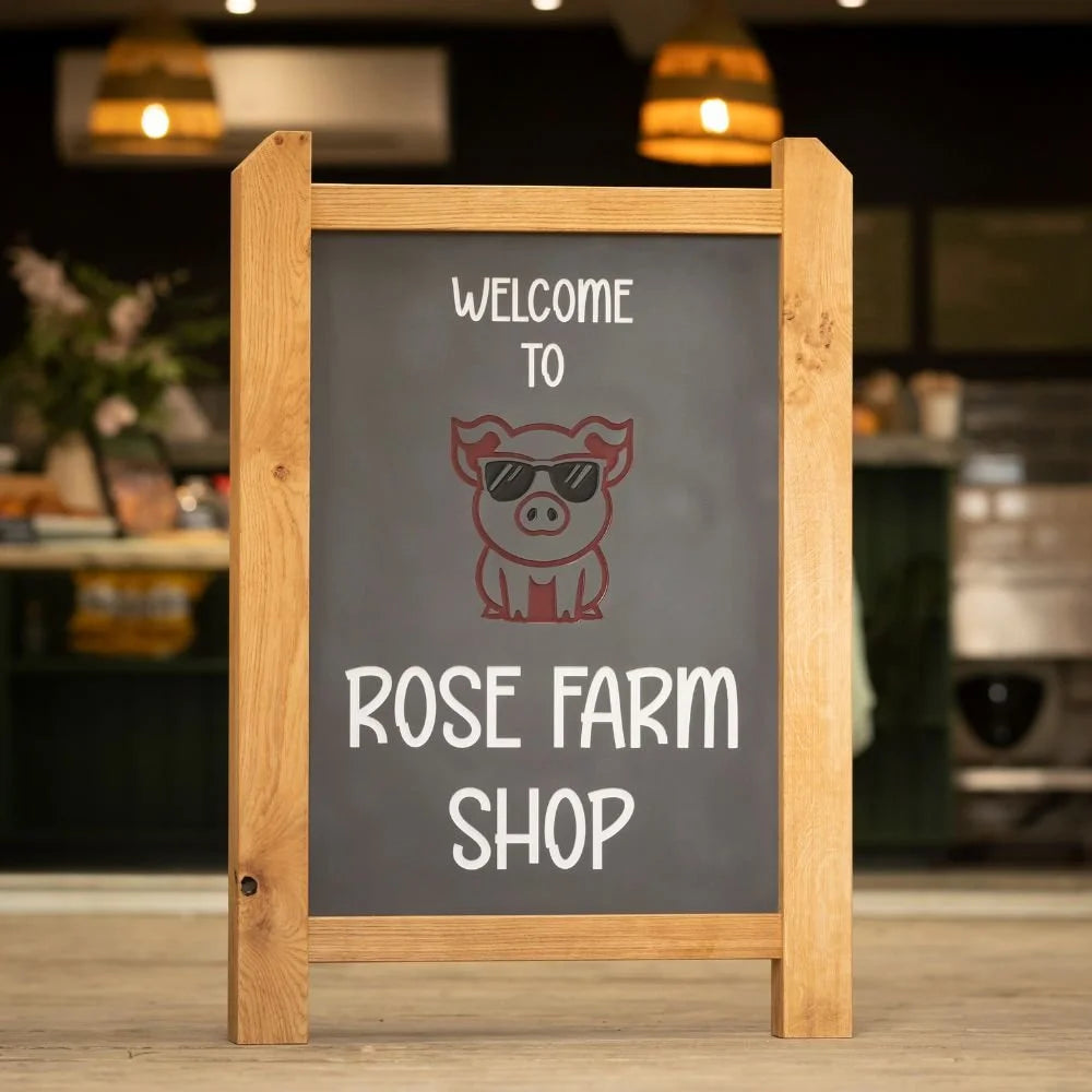 Chalkboard sign with 'Welcome to Rose Farm Shop' and a pig illustration in sunglasses on a blurred indoor background.