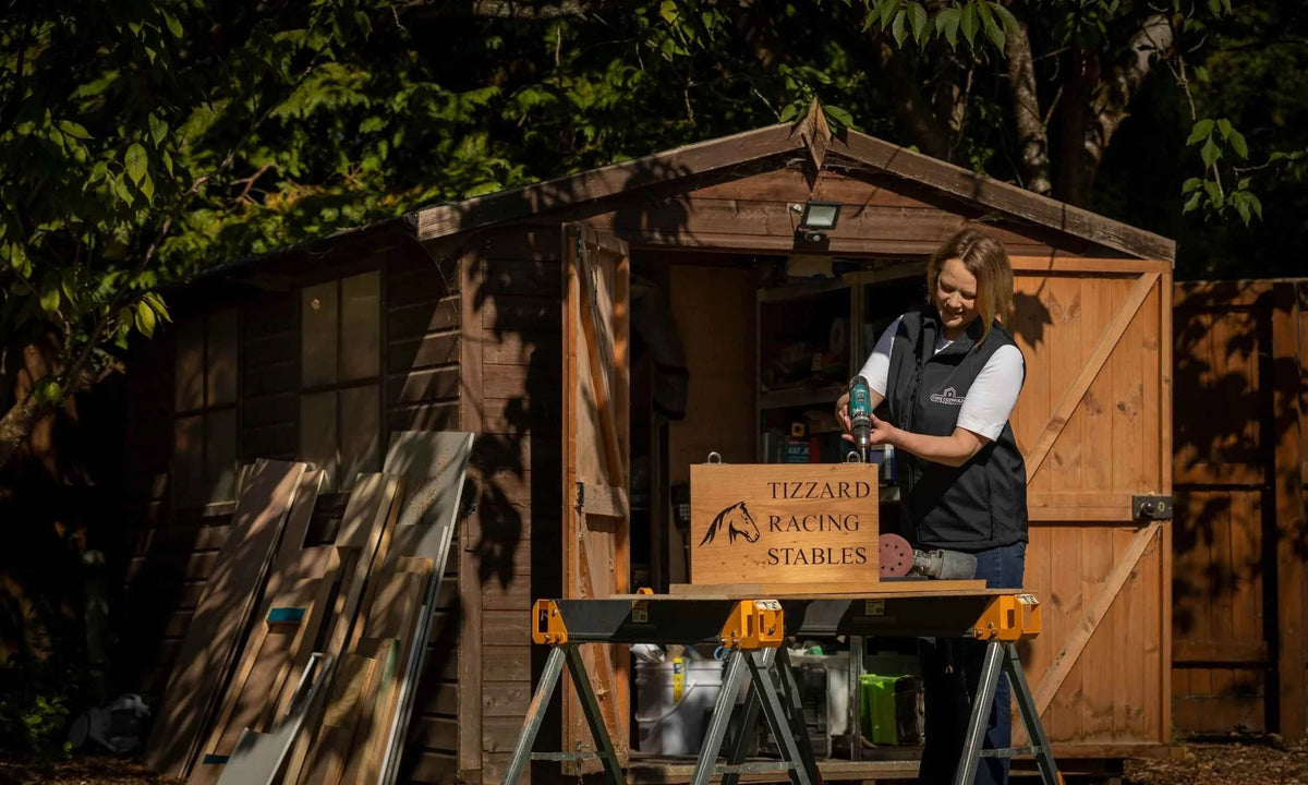 Person standing next to a wooden shed with 'Tizzard Racing Stables' branding.