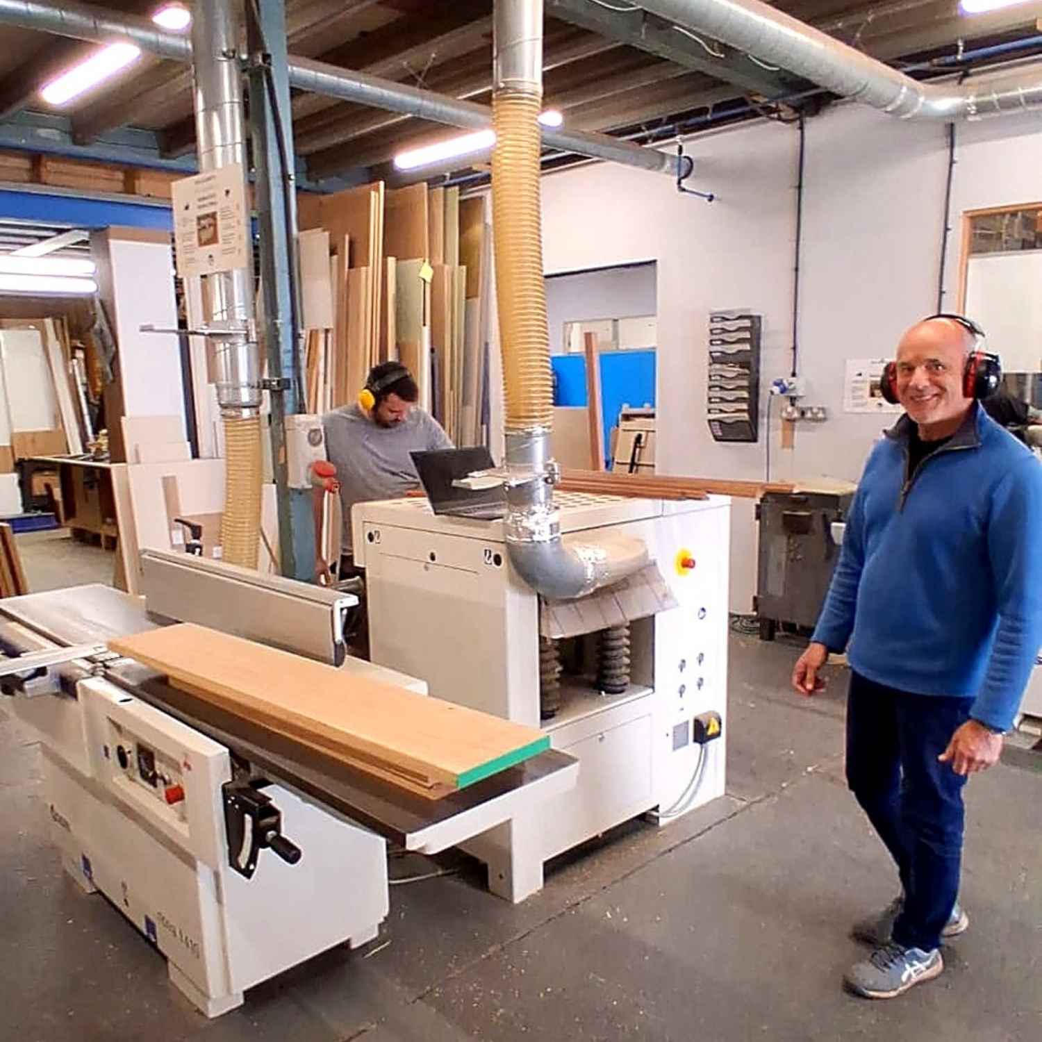 Man standing in a workshop with woodworking equipment and another person working in the background.