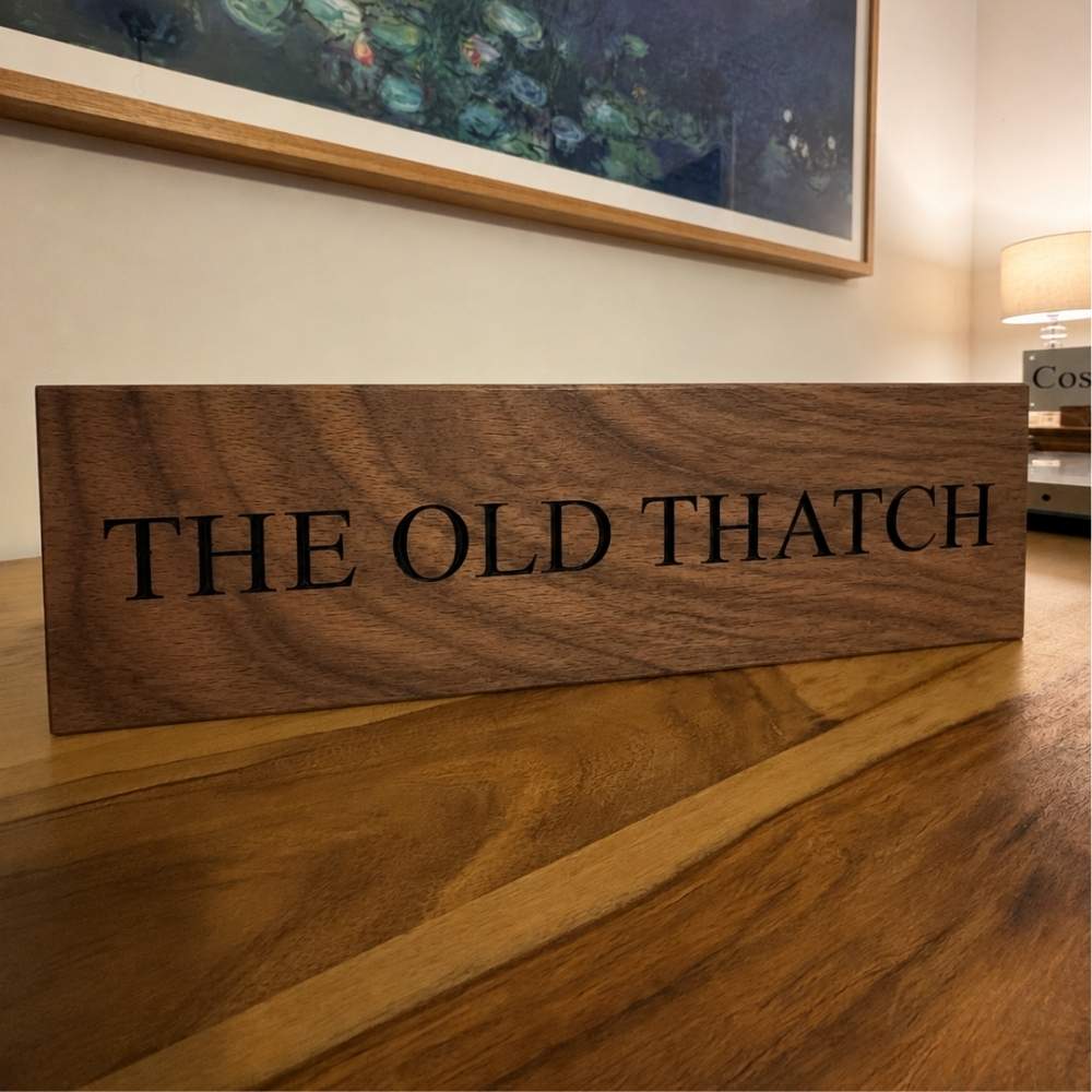 A horizontal walnut wooden sign engraved with 'THE OLD THATCH' in black serif letters, resting on a wooden table in a warmly lit room with a framed painting on the wall.