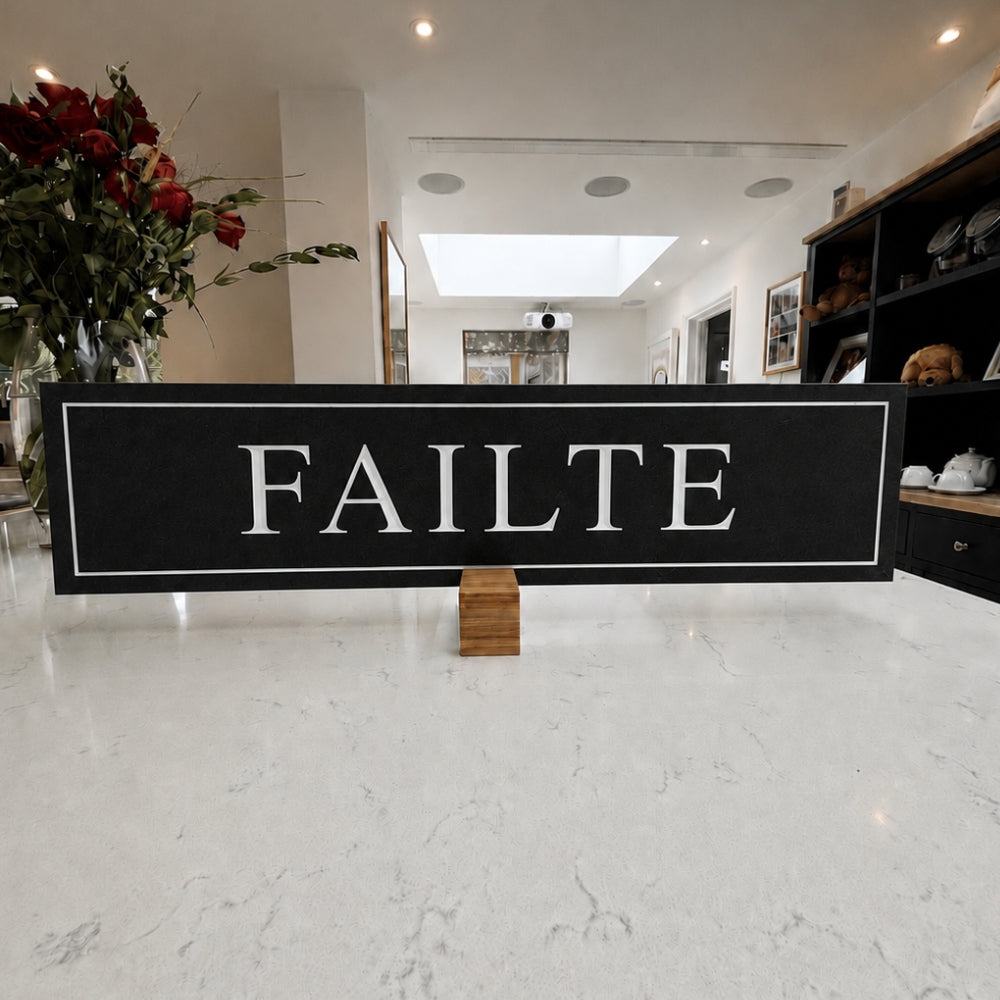 A rectangular black sign with the word 'FAILTE' in large white serif letters, displayed on a small wooden block in a bright modern interior with a white marble floor and decorative flowers in the background.