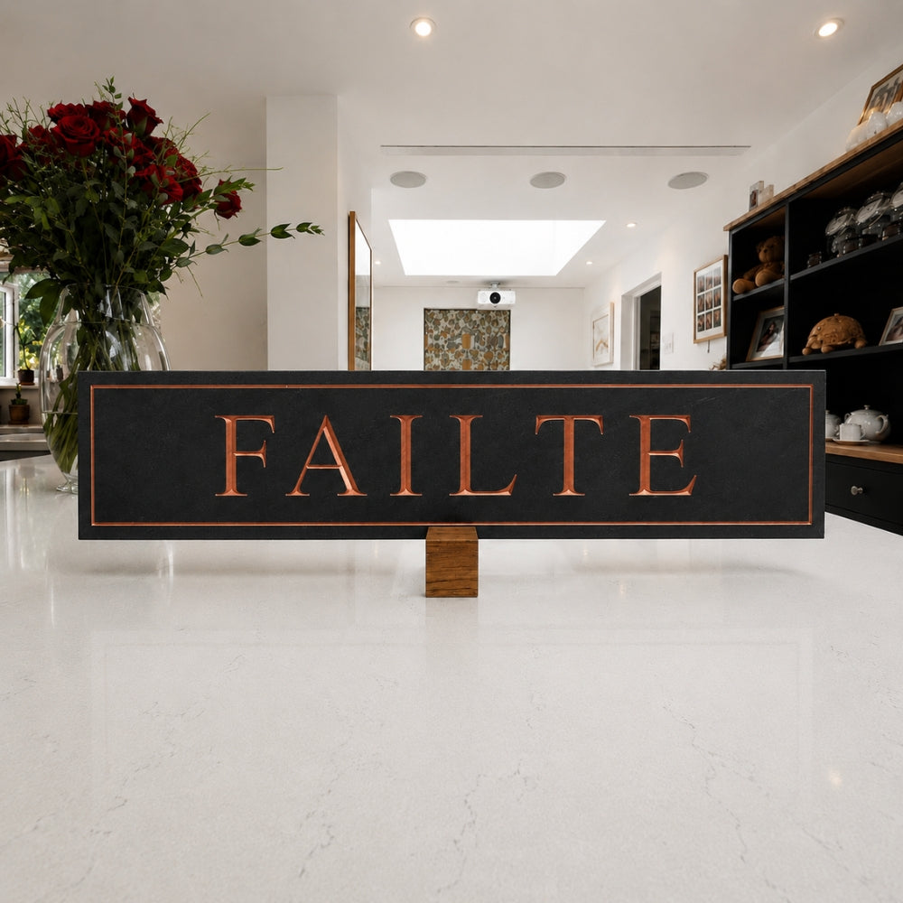 A rectangular black sign with the word 'FAILTE' in large copper-toned letters, displayed on a small wooden stand in a bright, modern interior hallway.