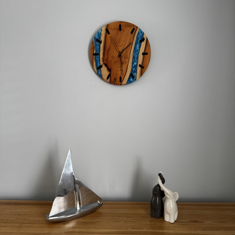 A round wall clock with a natural wood grain face and blue resin river details, mounted on a light gray wall above a wooden shelf with decorative objects.