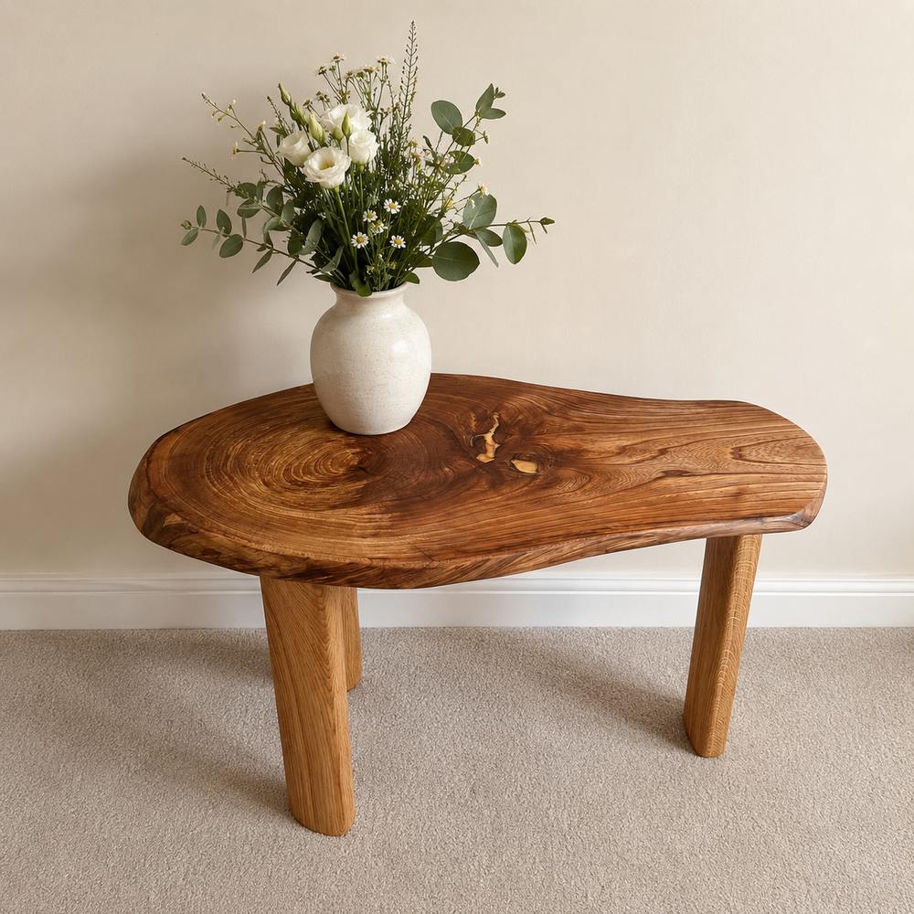 A live-edge wooden coffee table with a rustic natural wood grain, styled with a white ceramic vase of white flowers and green foliage, set against a neutral beige wall and carpet.