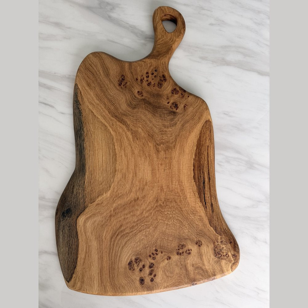A rustic live-edge wooden cutting board with a natural burl wood pattern and a hanging loop, displayed on a white marble surface.