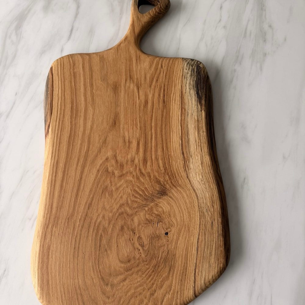 A rustic live-edge wooden cutting board with a natural handle, displayed on a white marble surface.