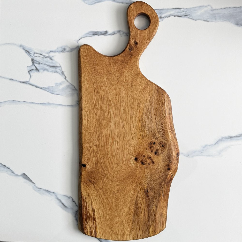 A rustic wooden serving board with a handle and a hole, displayed on a white marble surface with gray veining.