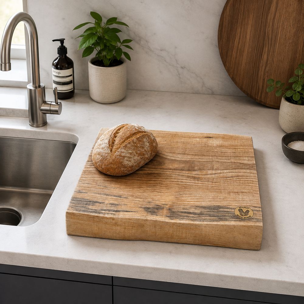 A rustic live-edge wooden cutting board with a small engraved heart logo, placed on a white marble kitchen countertop next to a sink. A round loaf of bread sits on the board, with potted plants and a soap dispenser in the background.