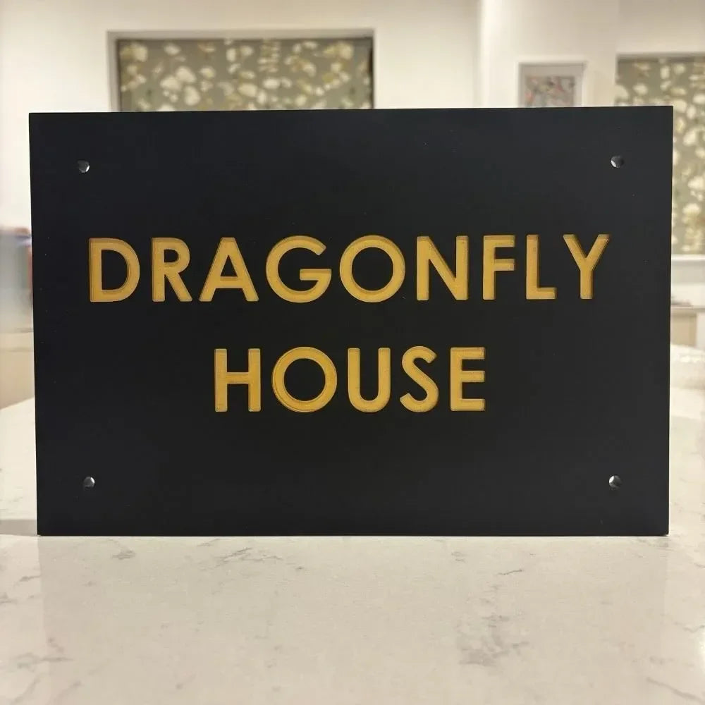 Black sign with 'DRAGONFLY HOUSE' in gold letters on a marble surface.