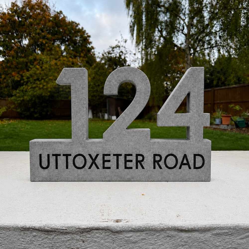 A granite house number sign displaying '124 UTTOXETER ROAD' in black lettering, set outdoors on a white wall with a blurred garden background.