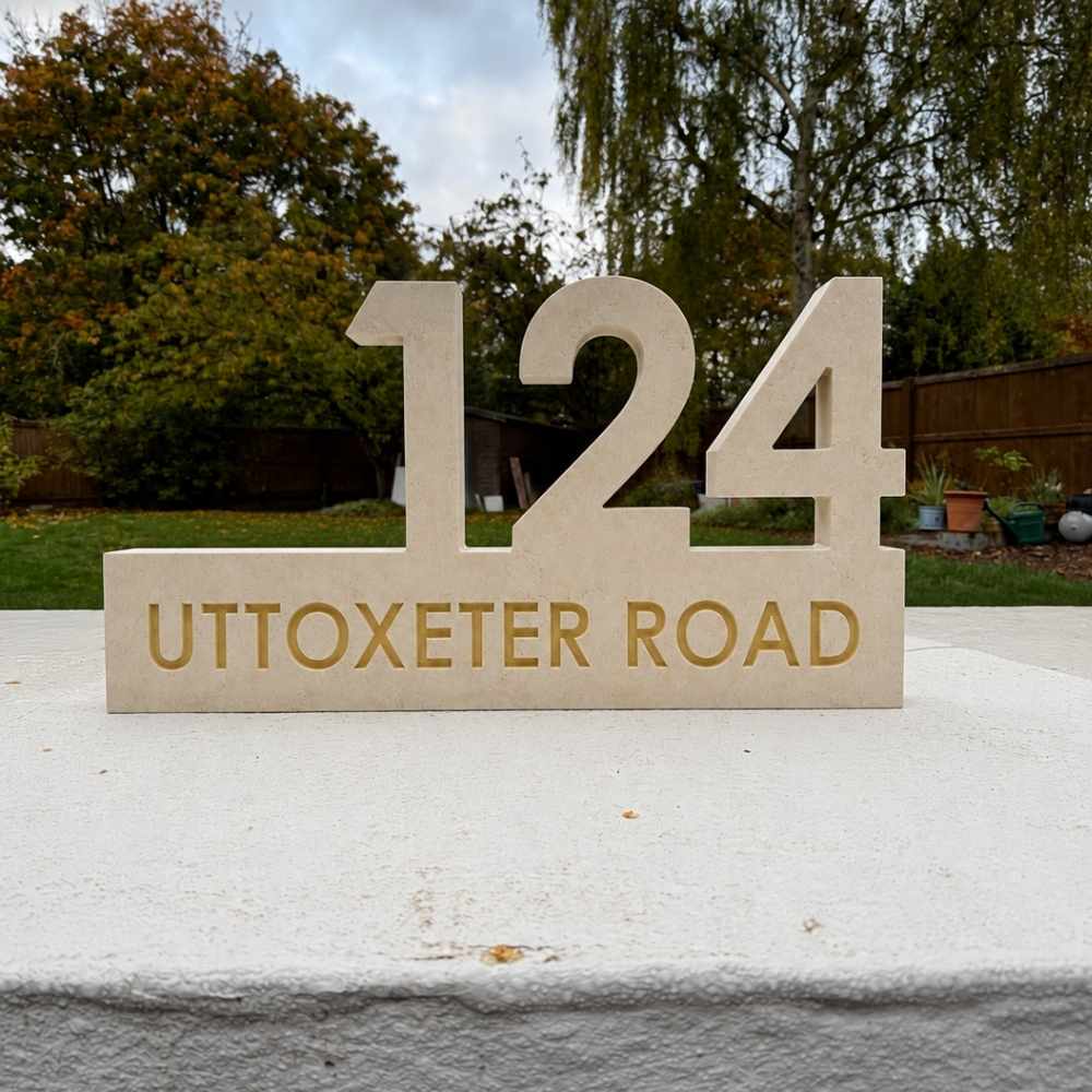 A freestanding limestone house number and road name sig with gold lettering, photographed outdoors on a white wall with trees and a fence in the background.