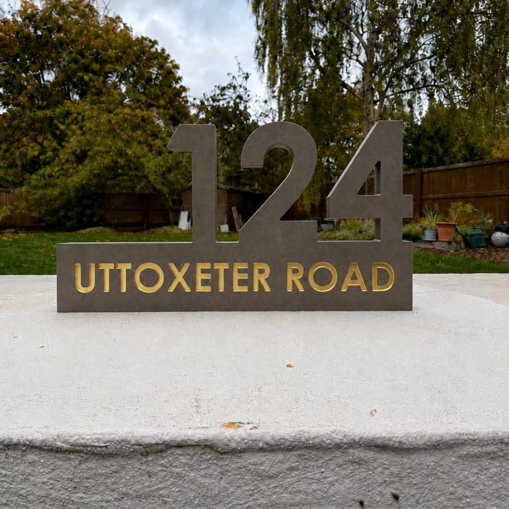 A large mink house number sign reading 124 UTTOXETER ROAD, mounted on a concrete surface with a garden background.