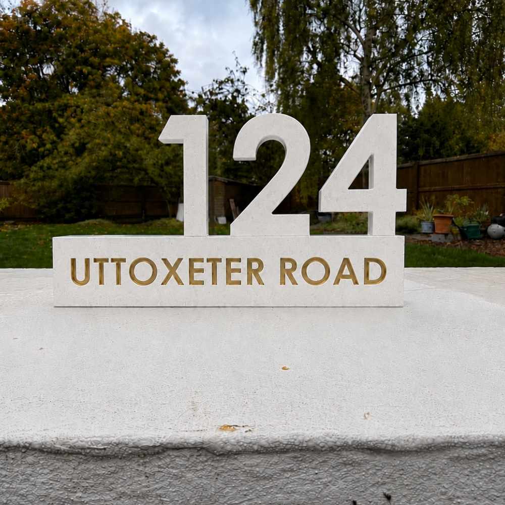 Large white freestanding house number sign reading 124 with gold lettering on a matching white sign that says UTTOXETER ROAD, placed outdoors on a concrete surface with trees and a fence in the background.