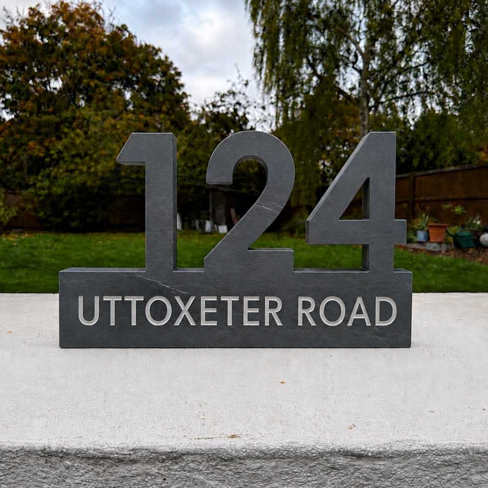A dark grey custom house number sign reading '124 UTTOXETER ROAD' mounted on a concrete wall with a garden background.