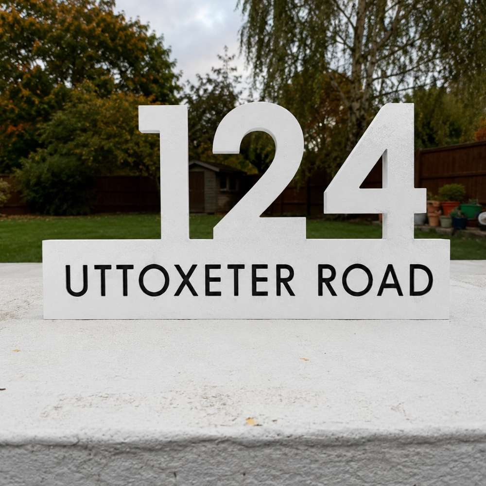 White freestanding house number sign reading 124 UTTOXETER ROAD, placed outdoors on a light-colored wall with trees and a garden in the background.