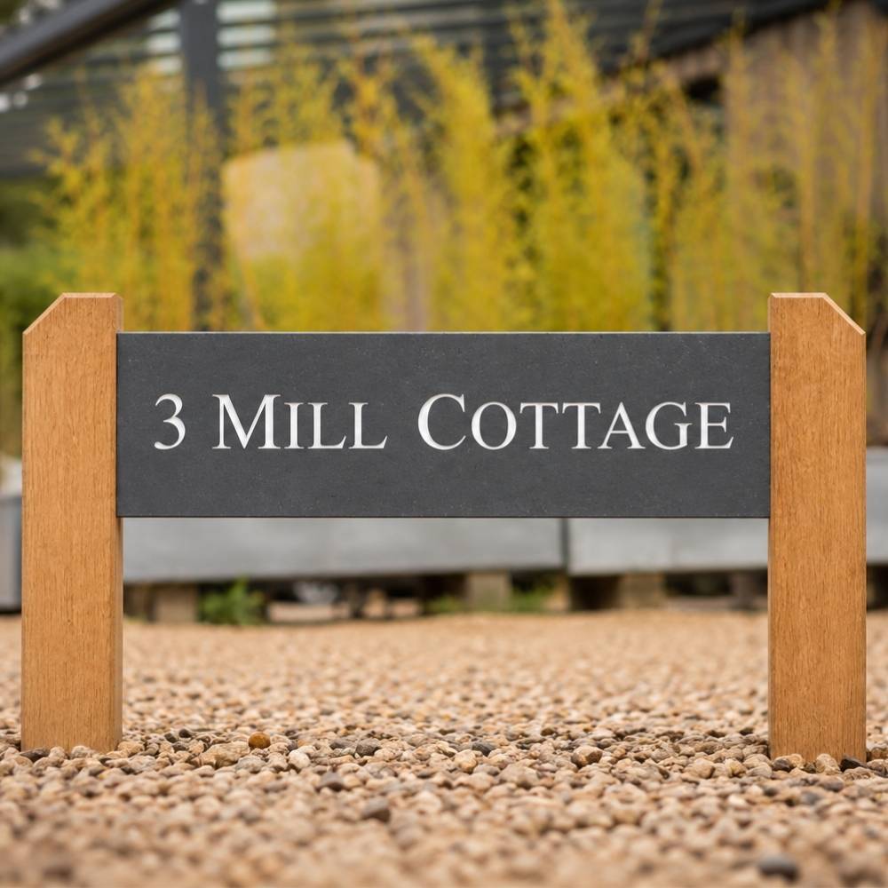 A custom engraved outdoor slate sign for '3 Mill Cottage'  with white lettering, supported by two light brown wooden posts on a gravel path.