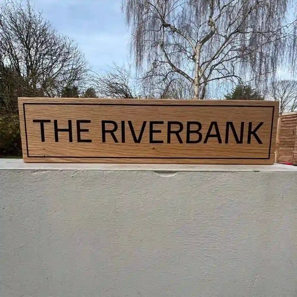Wooden sign with 'THE RIVERBANK' text on a wall with trees in the background