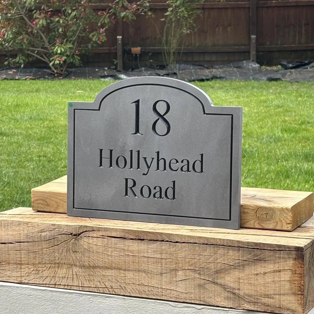 House number sign '18 Hollyhead Road' on a wooden base with grass and a wooden fence in the background.