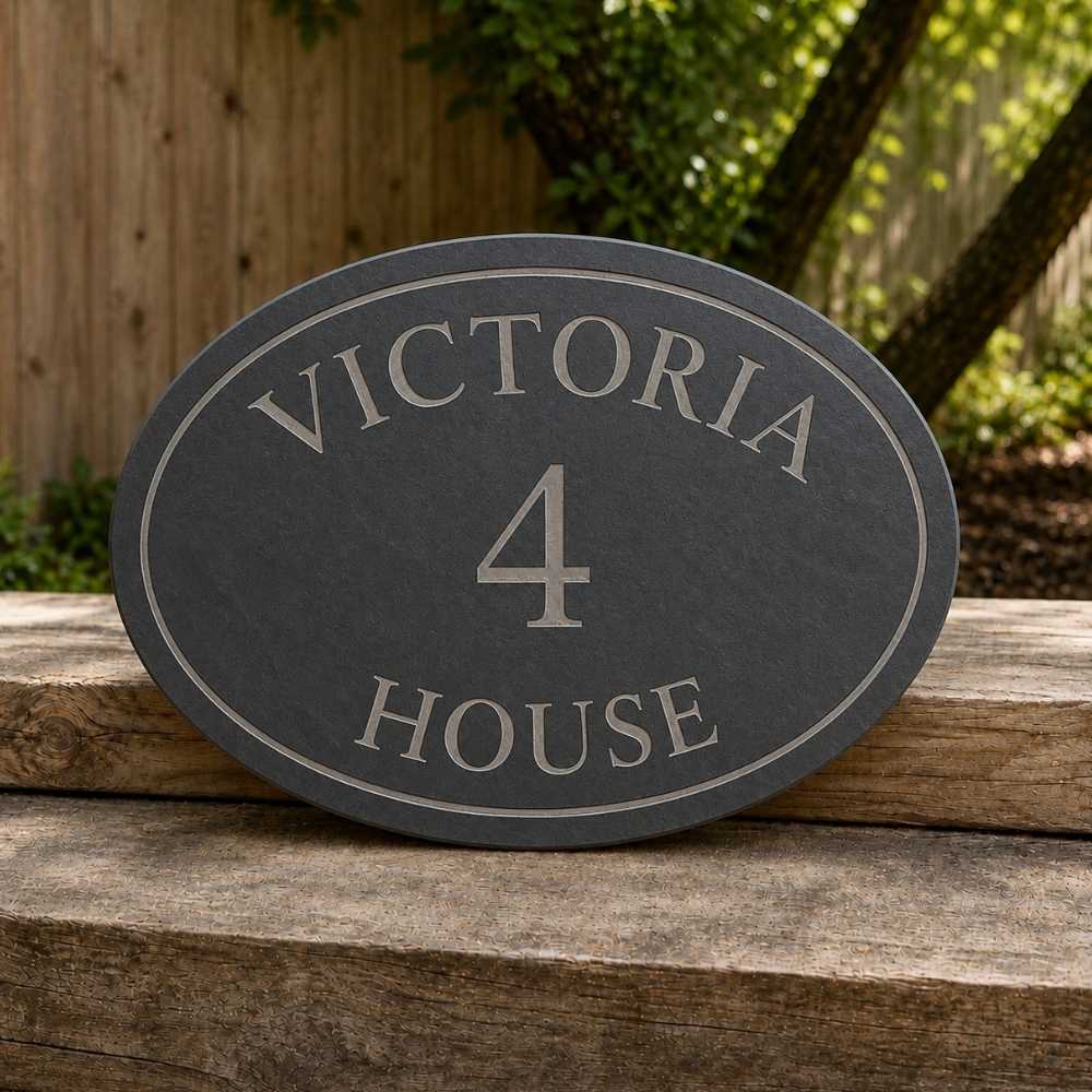 Oval dark slate house sign reading 'VICTORIA 4 HOUSE' in light gray lettering, displayed on a rustic wooden surface with a blurred garden background.