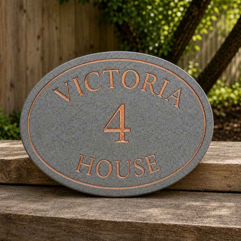 A charcoal house plaque with copper lettering reading 'VICTORIA 4 HOUSE', displayed outdoors on a wooden surface with a blurred fence and greenery in the background.