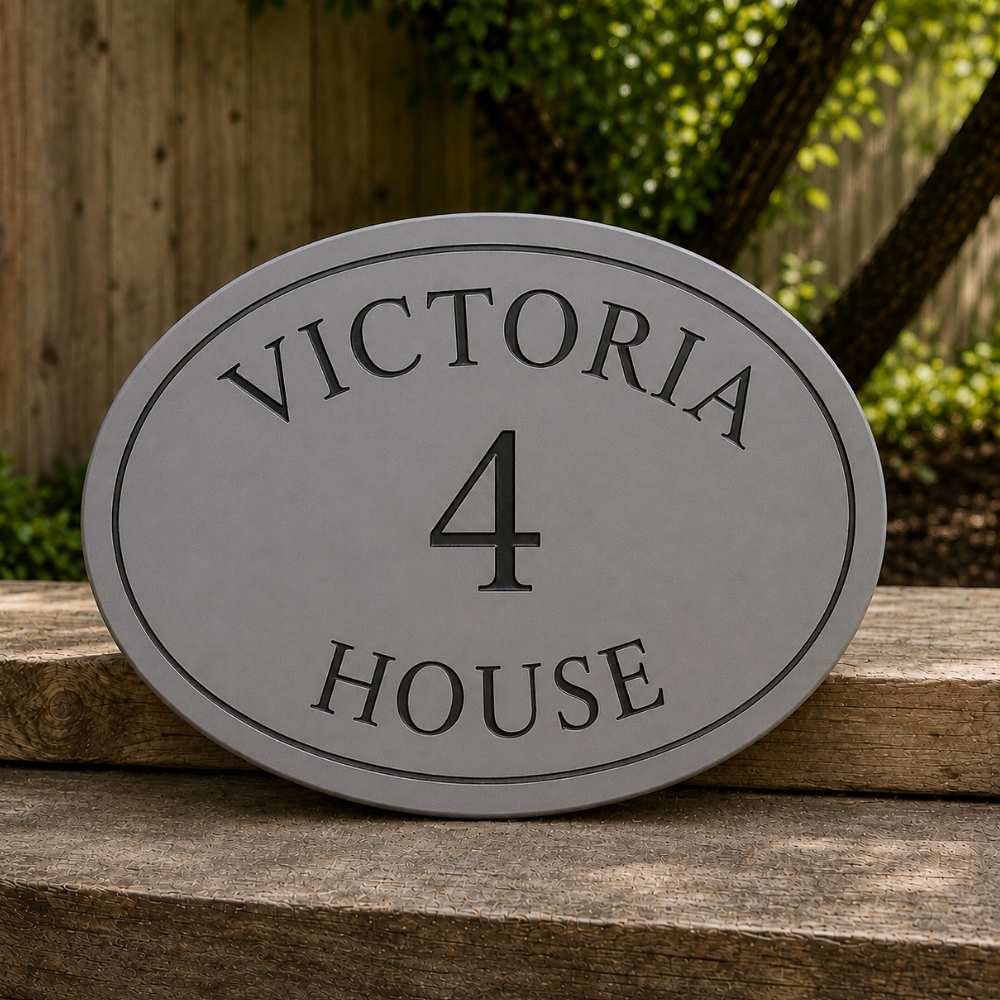 An oval grey house number sign reading 'VICTORIA 4 HOUSE' in black lettering, displayed outdoors on a weathered wooden surface with a blurred garden background.