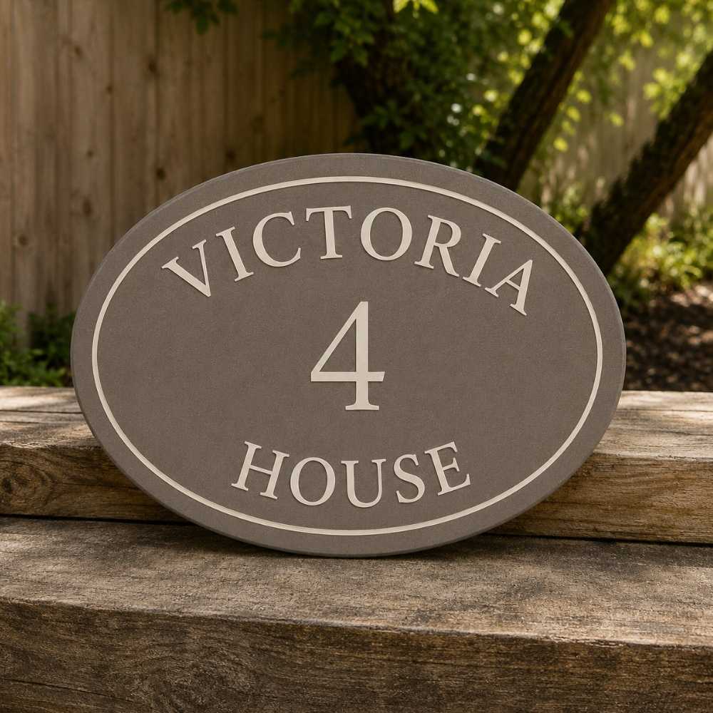 An oval mink house plaque reading 'VICTORIA 4 HOUSE' in light beige lettering on a dark gray background, displayed outdoors on a rustic wooden surface with a fence and greenery behind it.