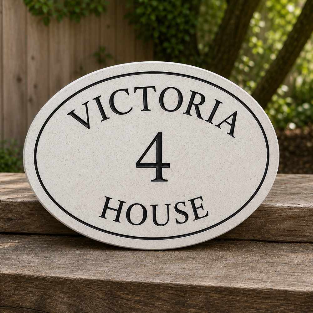 An oval quartz house number sign reading 'VICTORIA HOUSE 4' in black lettering on a light beige stone or concrete plaque, shown resting on a wooden surface outdoors with a fence and greenery in the background.