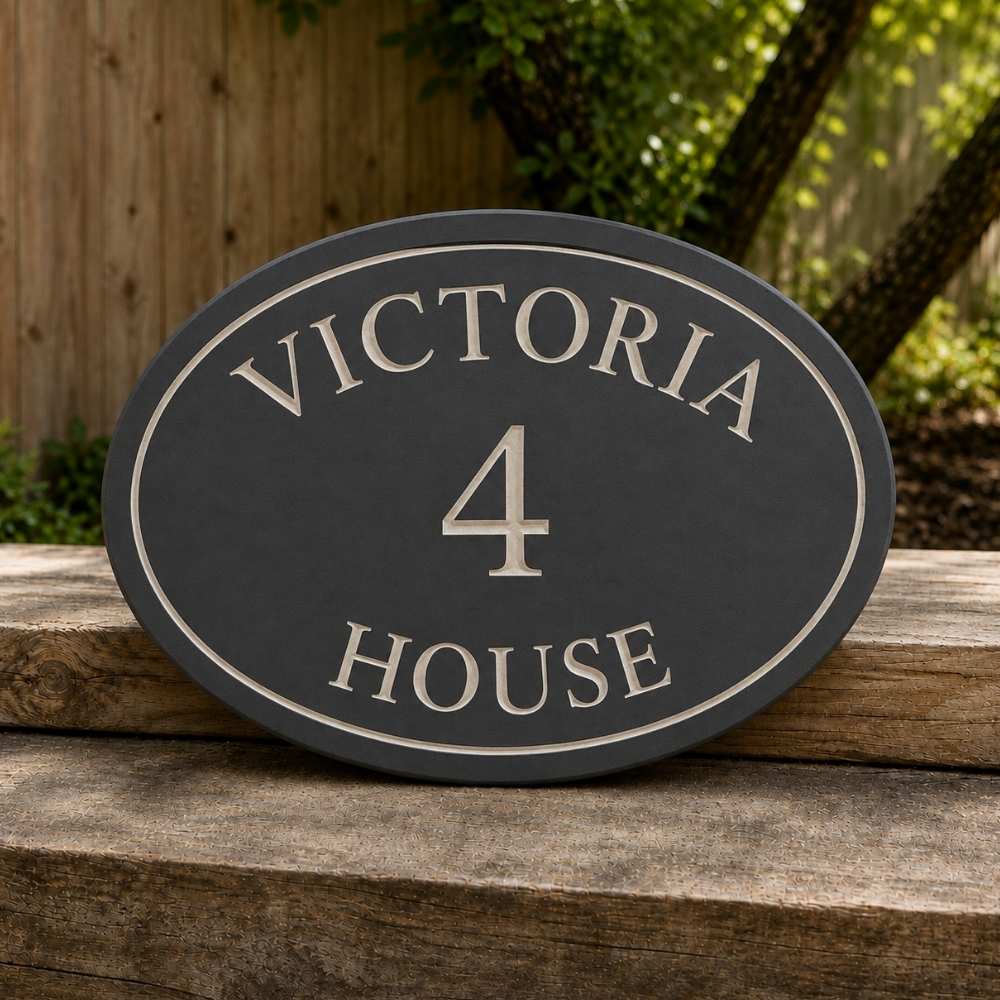 An oval  slate house plaque with raised cream lettering reading 'VICTORIA 4 HOUSE', displayed outdoors on a wooden surface with a garden fence and greenery in the background.