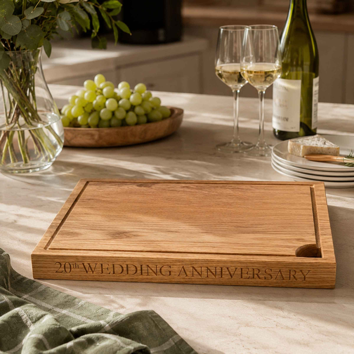 Wooden cutting board with '20th Wedding Anniversary' engraving on a table with wine and grapes.