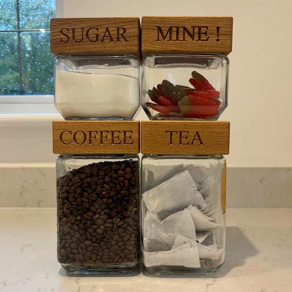 Four clear glass jars with wooden lids labeled SUGAR, MINE!, COFFEE, and TEA, arranged on a countertop. The jars contain white sugar, red peppers, coffee beans, and tea bags.