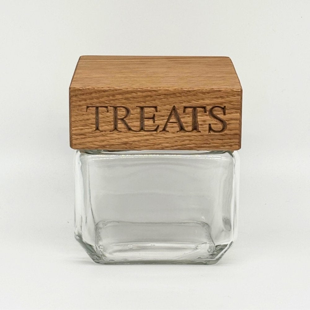A clear glass jar with a square wooden lid engraved with the word 'TREATS' in dark lettering, shown on a plain light gray background.