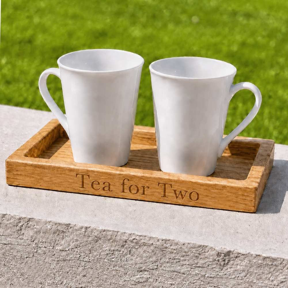 Two white ceramic mugs sitting in a rectangular wooden tray engraved with the words 'Sunday Mornings', placed outdoors on a stone ledge with green grass in the background.