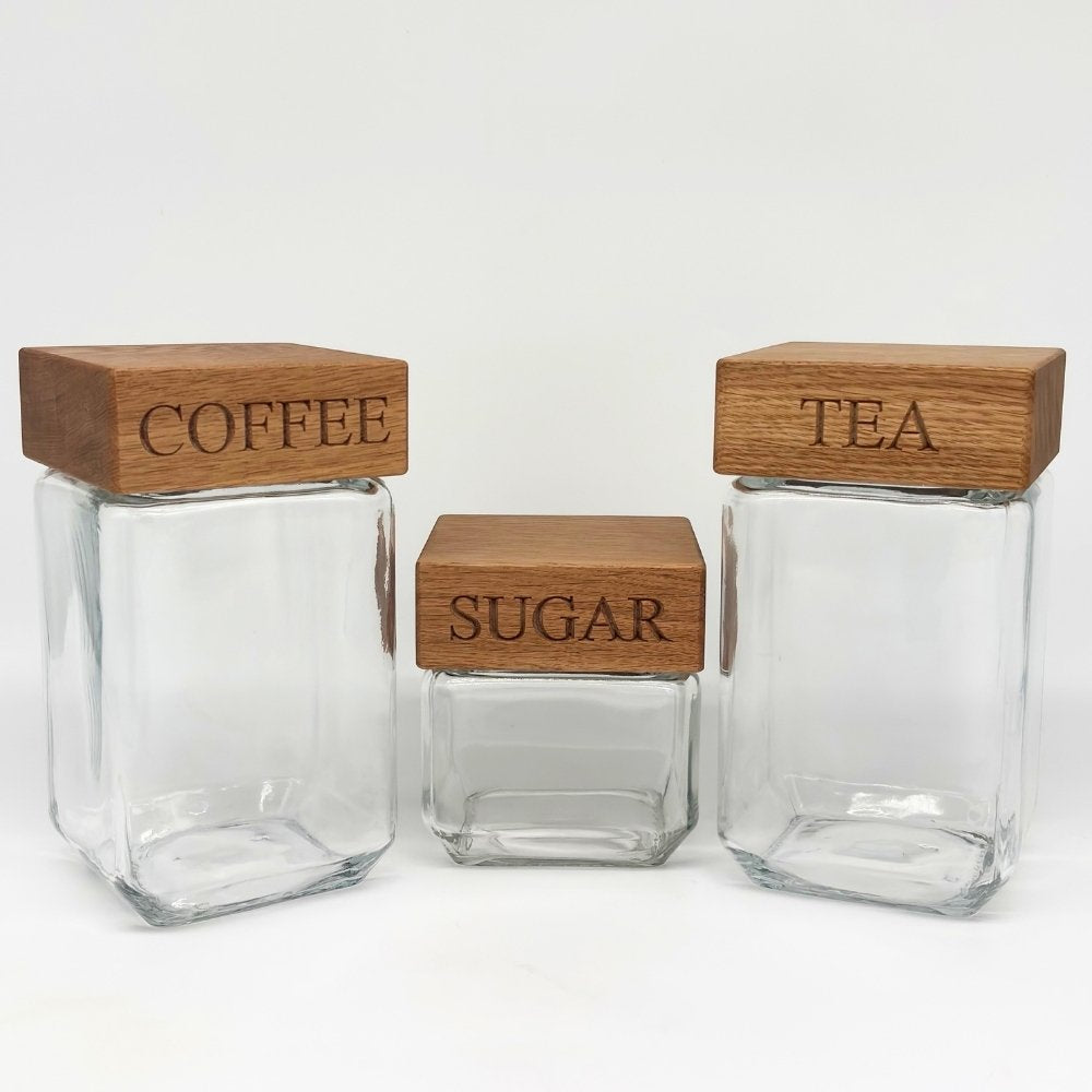 Three clear glass canisters with rectangular wooden lids labeled COFFEE, SUGAR, and TEA, arranged on a white studio background.