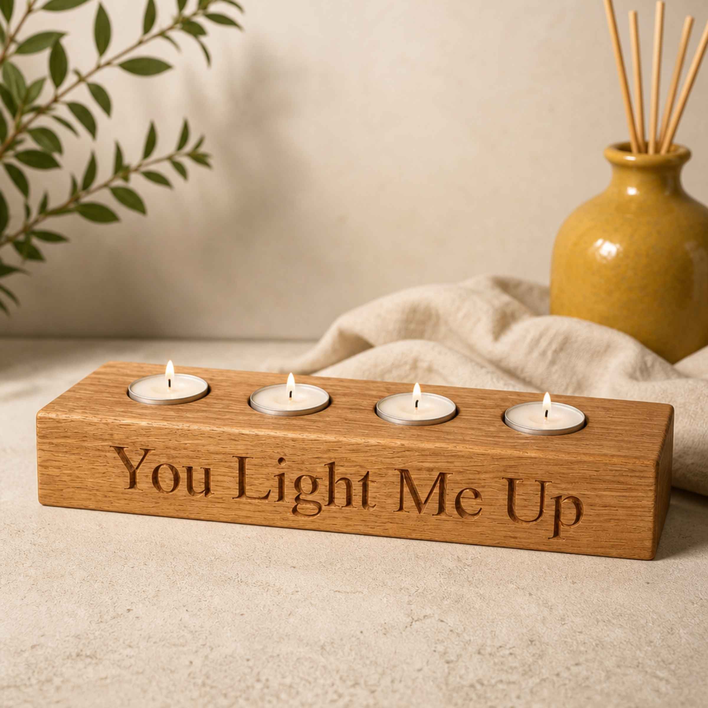 Wooden candle holder with four tealights and 'You Light Me Up' engraving, placed on a neutral surface with a vase and plant in the background.