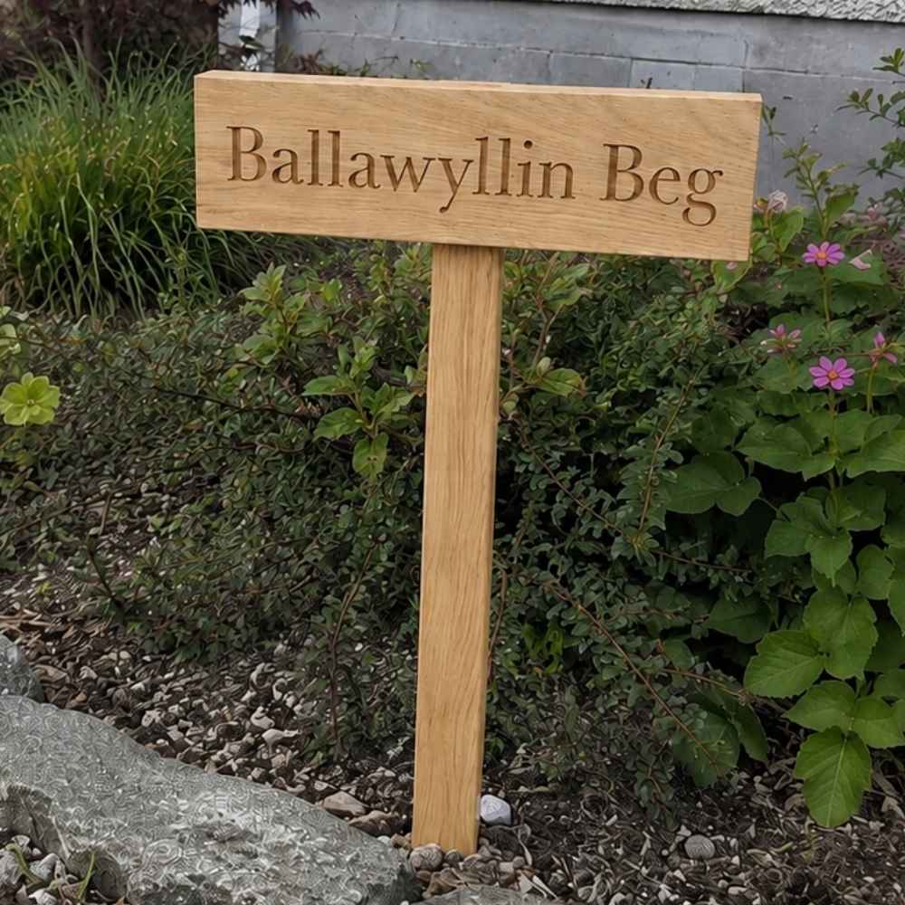A rectangular wooden sign engraved with 'Ballawyllin Beg' mounted on a single wooden stake, set in a garden bed with green plants and gravel.
