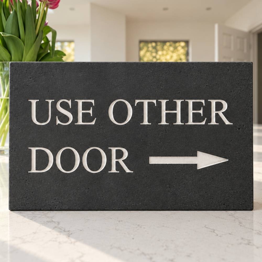 A rectangular dark slate stone with off white (pebble) text reading 'USE OTHER DOOR' and a white arrow pointing right, placed on a light marble surface in a bright indoor setting.