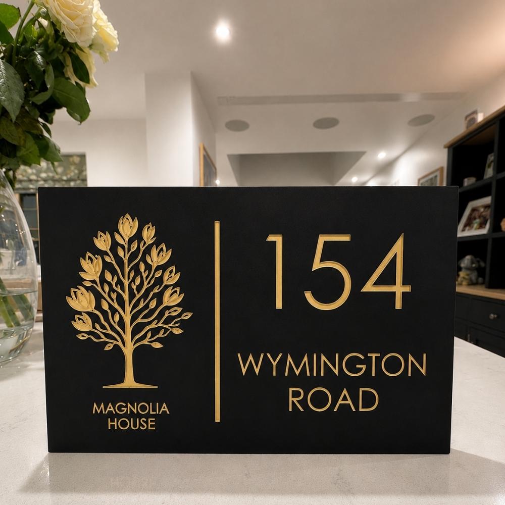 A black rectangular house sign with gold lettering and a gold tree emblem, reading '154 WYMINGTON ROAD' and 'MAGNOLIA HOUSE,' placed on a countertop in a home interior.