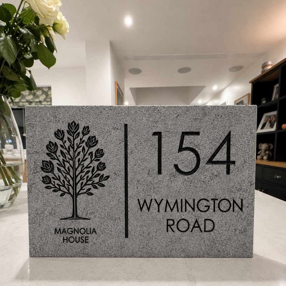 A rectangular granite stone house number plaque on a countertop, featuring a black engraved tree logo and the text 'MAGNOLIA HOUSE' alongside '154 WYMINGTON ROAD'. The background shows a blurred interior room with flowers on the left and shelving on the right.