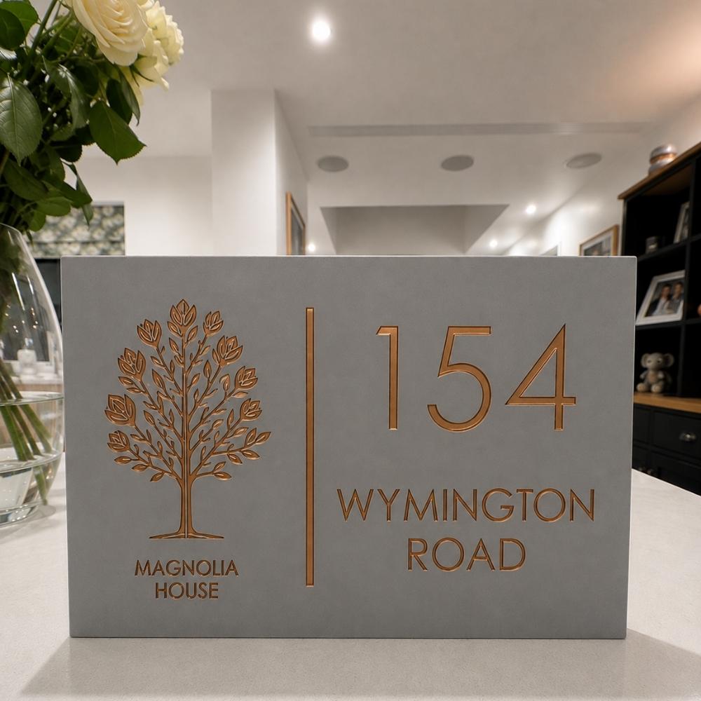 A rectangular grey house number sign with a gold tree emblem and the text '154 WYMINGTON ROAD' and 'MAGNOLIA HOUSE', displayed on a countertop in a home interior.