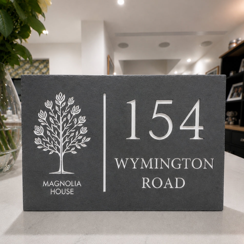A rectangular slate house number plaque with a white tree logo and the text 'MAGNOLIA HOUSE' and '154 WYMINGTON ROAD', displayed on a light countertop in a home interior setting.