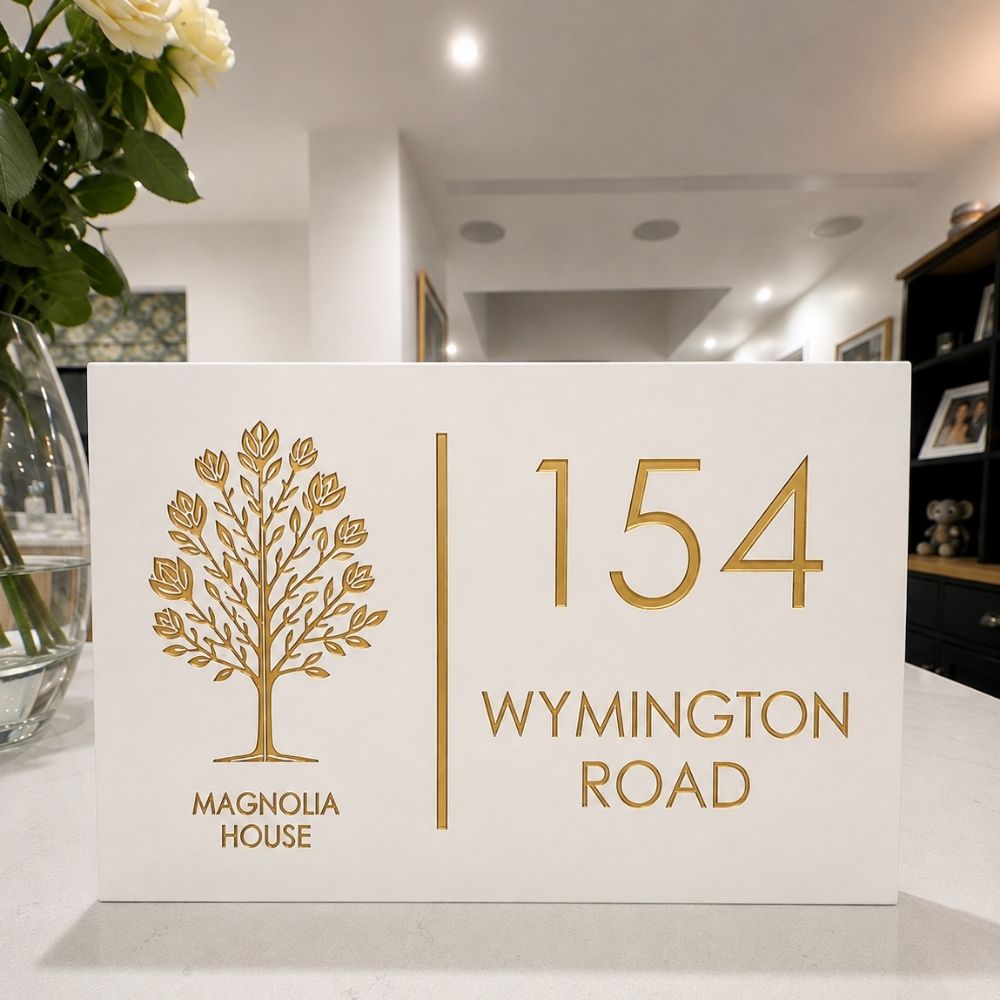 A white house number sign with gold lettering and a gold tree emblem, reading '154 WYMINGTON ROAD' and 'MAGNOLIA HOUSE,' placed on a countertop in a home interior.