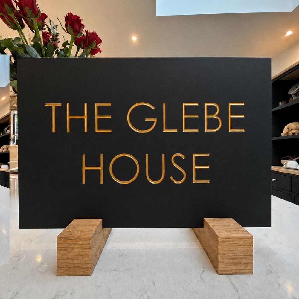A black rectangular sign with raised gold lettering reading 'THE GLEBE HOUSE' displayed on a white marble counter in a modern interior setting, supported by two wooden blocks.