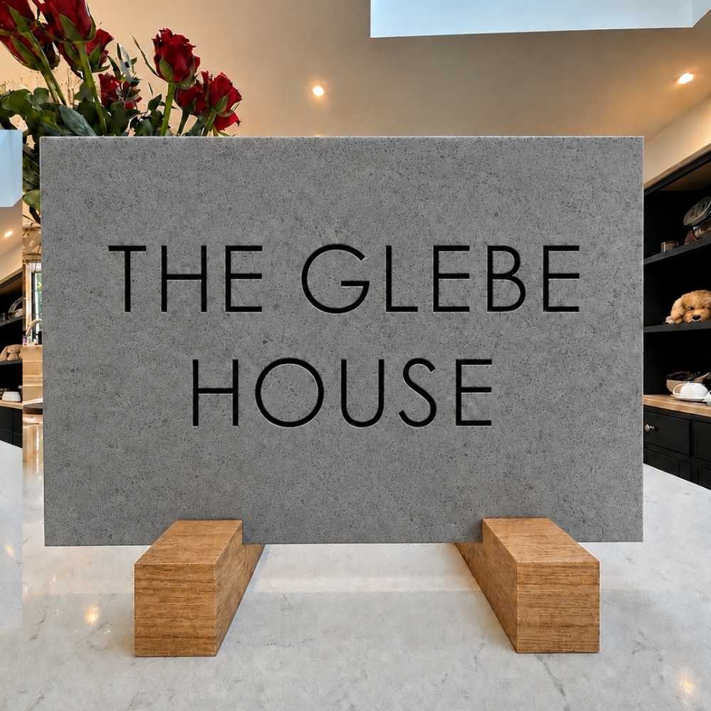 A rectangular charcoal stone sign with the words 'THE GLEBE HOUSE' in black serif lettering, displayed on two wooden blocks on a light marble surface in an indoor setting.