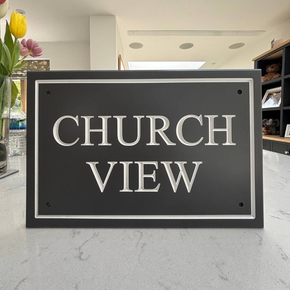 A rectangular dark slate house sign with raised silver lettering that reads 'CHURCH VIEW', photographed on a light marble countertop in a real indoor setting with flowers and shelving visible in the background.