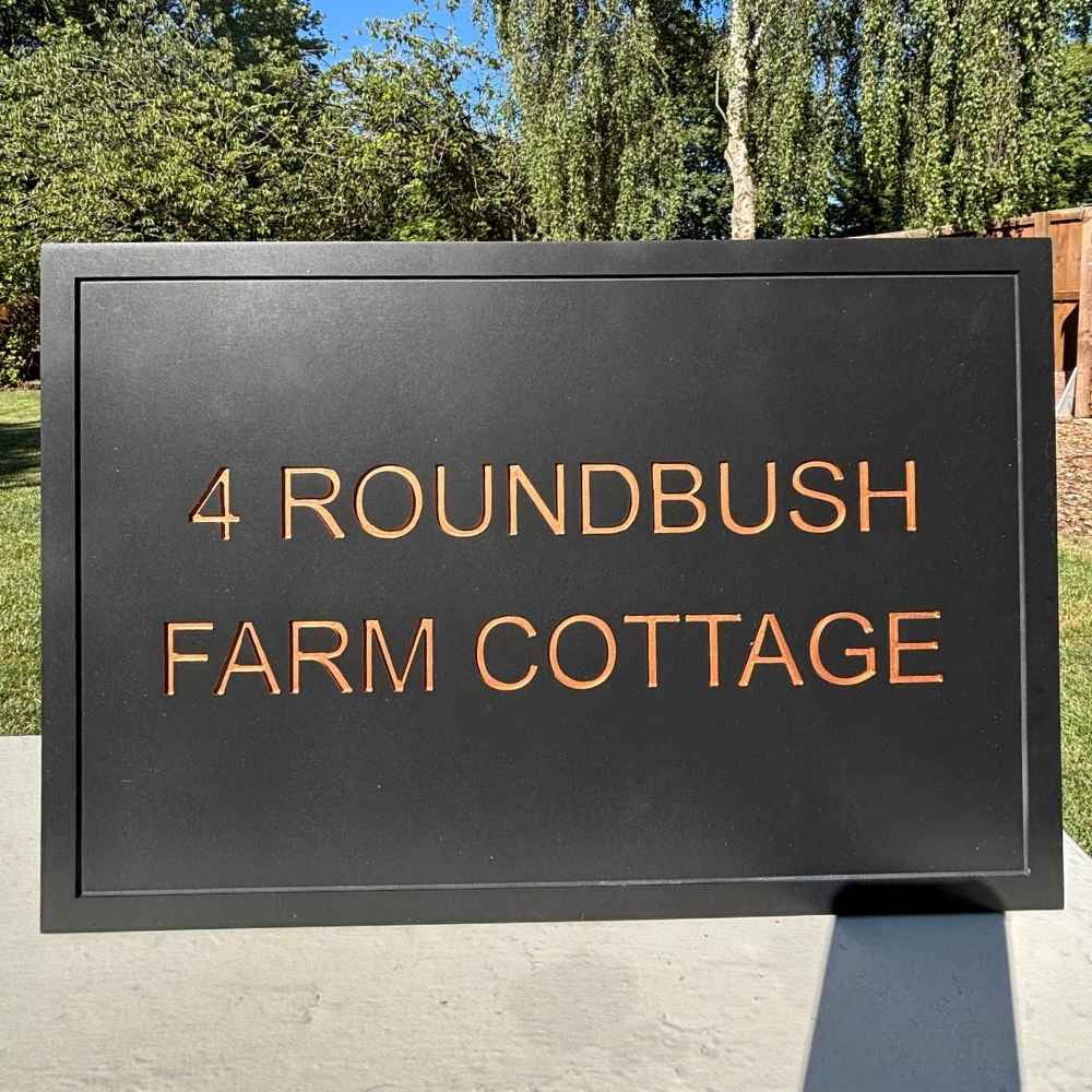 A rectangular black stone sign with raised copper-toned lettering reading '4 ROUNDBUSH FARM COTTAGE,' photographed outdoors against trees and grass.