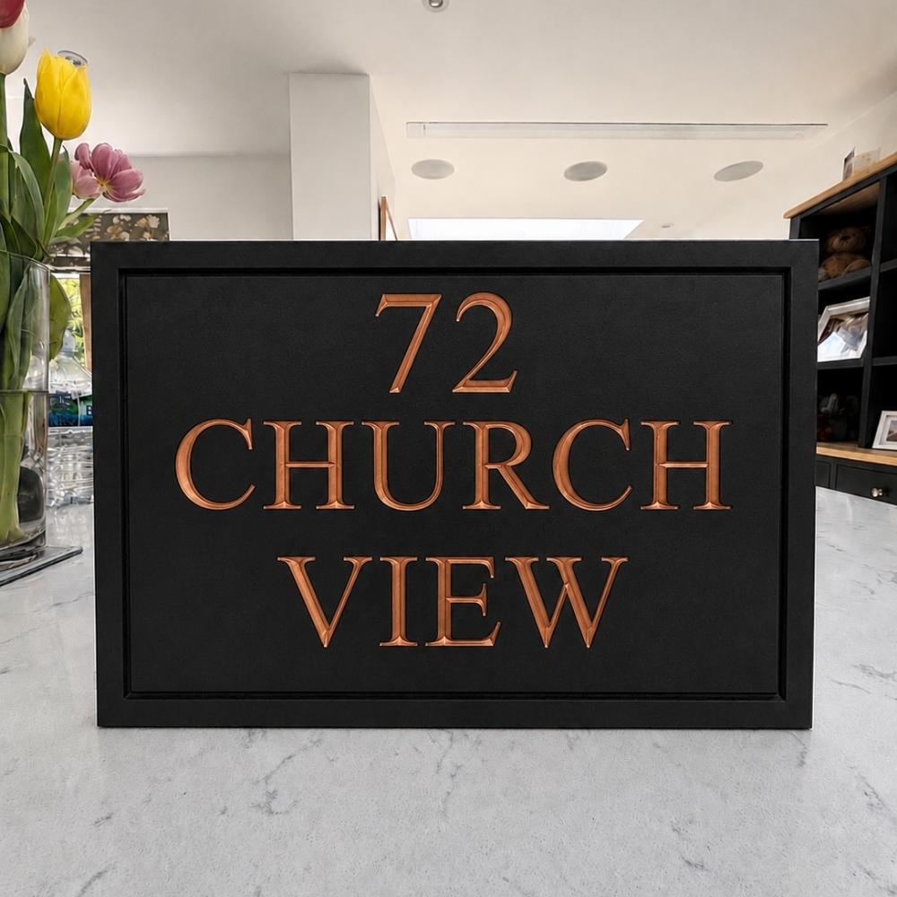 A black rectangular house number sign with raised copper-toned lettering reading '72 CHURCH VIEW', displayed on a light marble countertop in a real indoor setting with flowers and shelving visible in the background.