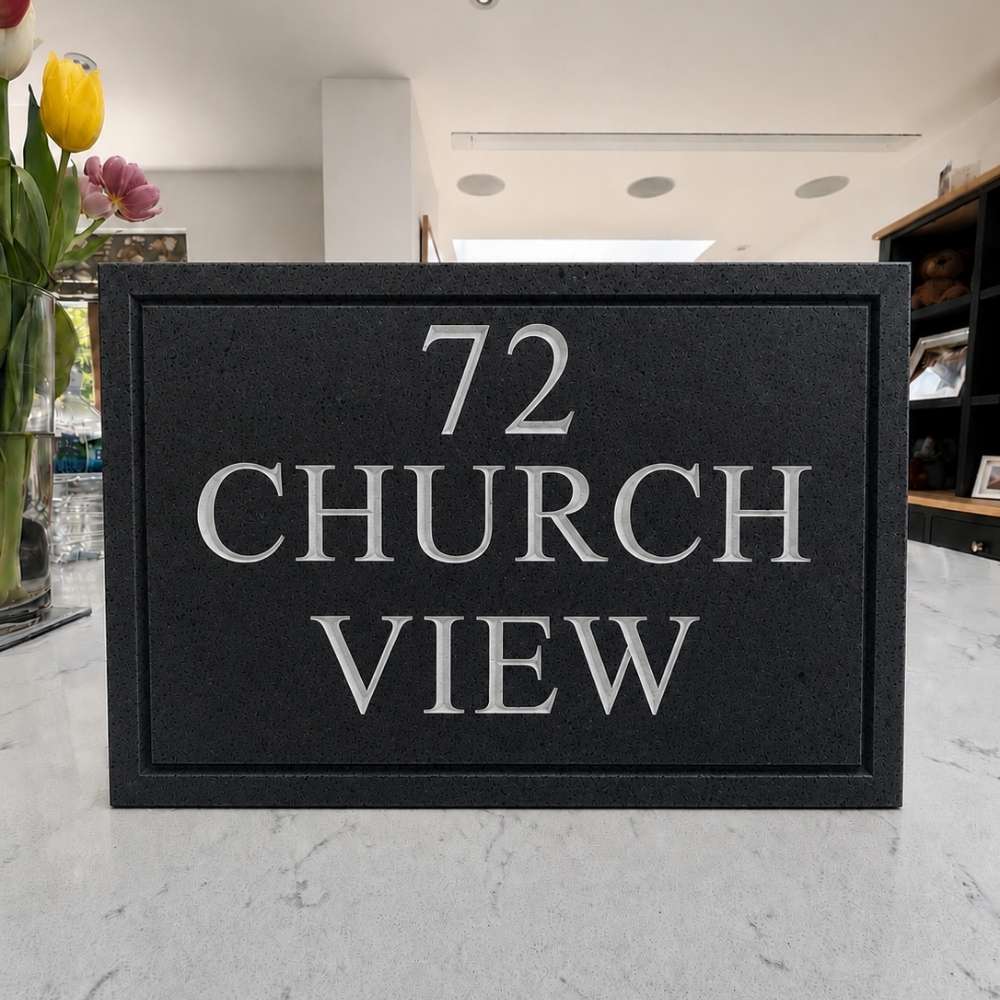A rectangular charcoal house plaque with raised silver lettering reading '72 CHURCH VIEW' on a light marble countertop, with flowers and a home interior visible in the background.